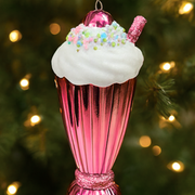 Pink ice cream cone ornament with whipped cream and sprinkles against a blurred Christmas tree background.
