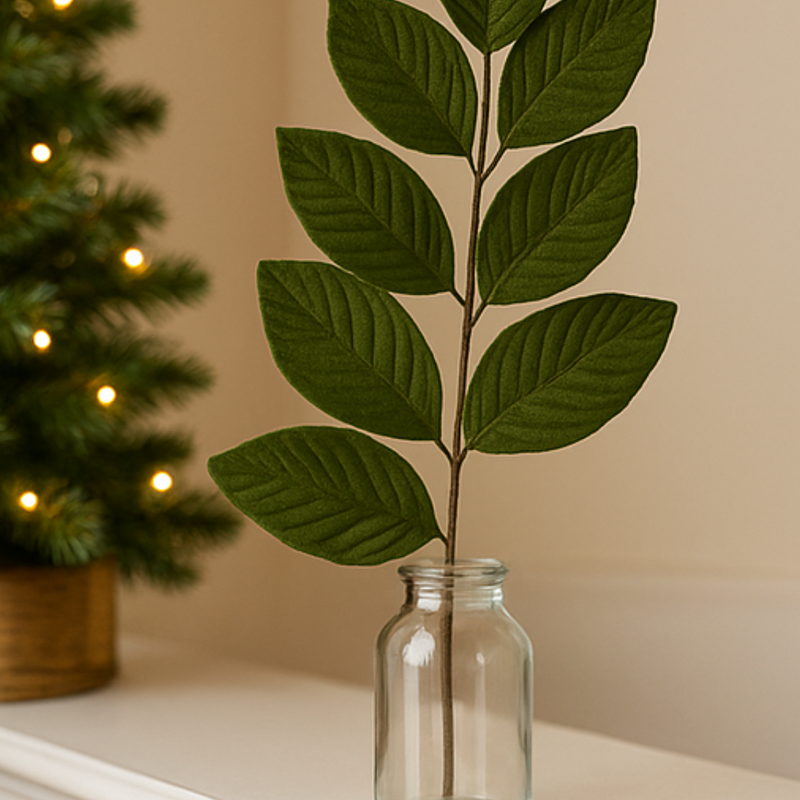 Green leaf branch in a clear glass vase against a beige wall with a blurred Christmas tree in the background.