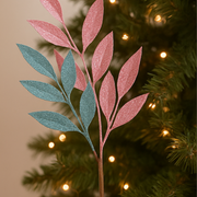 Decorative leaf branch with pink and blue leaves against a Christmas tree background.