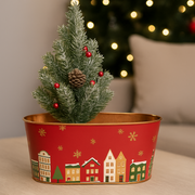 Small decorated Christmas tree in a red tin with festive village design, blurred Christmas tree in the background.