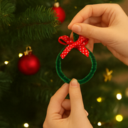 Hand holding a green ornament with a red bow in front of a decorated Christmas tree.