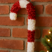 Decorative candy cane against a brick wall with a Christmas tree in the corner.